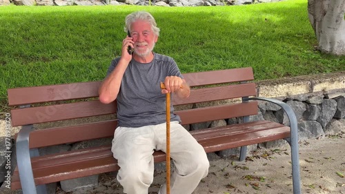 Happy cheerful elder 70s man sitting in a bench in the park talking on phone, senior smiling bearded grandfather enjoying free time and nature in outdoors