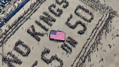 San Francisco, United States - 25 October 2025: Aerial view of crowds forming the words 'NO KINGS YES US 50' around a large American flag on a sandy surface.