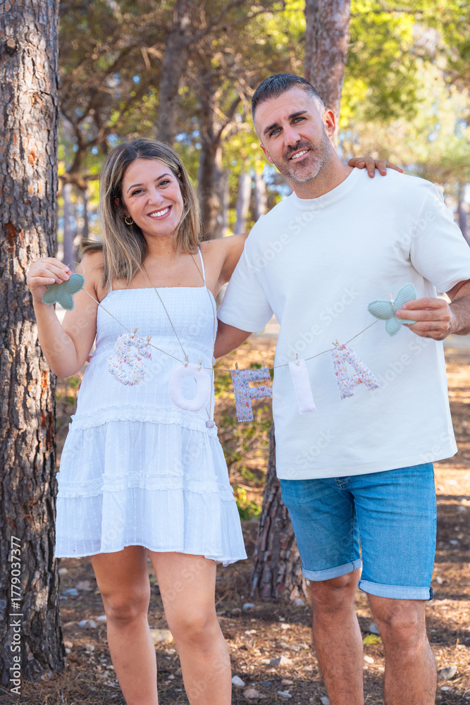 Naklejka premium Pregnant couple holding baby name letters outdoors in sunny nature. Future parents smiling with happiness and tenderness, celebrating pregnancy and family love in warm bright light.Vertical