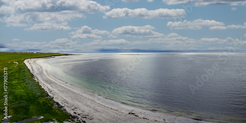 Fotografie Aerial view shows pale sand curving along a calm steel blue bay in Iceland's north, with green pastures, a faint track, and turquoise shallows in diffuse daylight