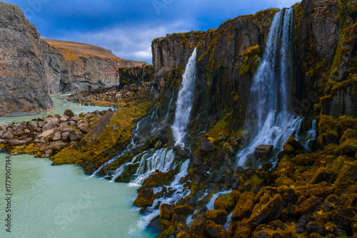 Water pours over dark basalt into turquoise water, mossy rocks and rugged canyon walls frame the view in Iceland. Soft overcast light enhances texture and contrast.
