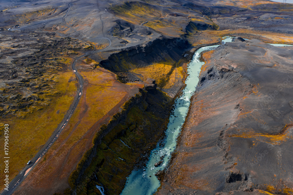 Fototapeta premium Aerial view shows Sigoldugljufur in Icelands Highlands, a milky turquoise river in a deep canyon, basalt and ash banks, golden moss, a gravel road with vehicles, autumn light.