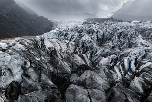 Foto Aerial view shows a jagged glacier in Vatnajokull National Park, Iceland, crevassed ice with dark ash, distant mountains, and a milky glacial lagoon at left in cool light