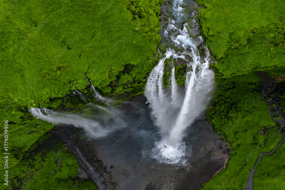 Fototapeta premium Aerial view shows Seljalandsfoss in southern Iceland as water drops from a mossy cliff into a dark pool, streams fan out in white ribbons, mist rises in daylight.