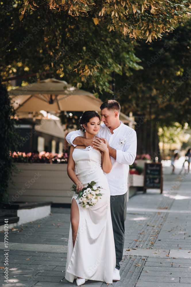 Naklejka premium Wedding photography, portrait of happy smiling newlyweds, stylish groom and beautiful young brunette bride, hugging outdoors on the street in the city, in the park.