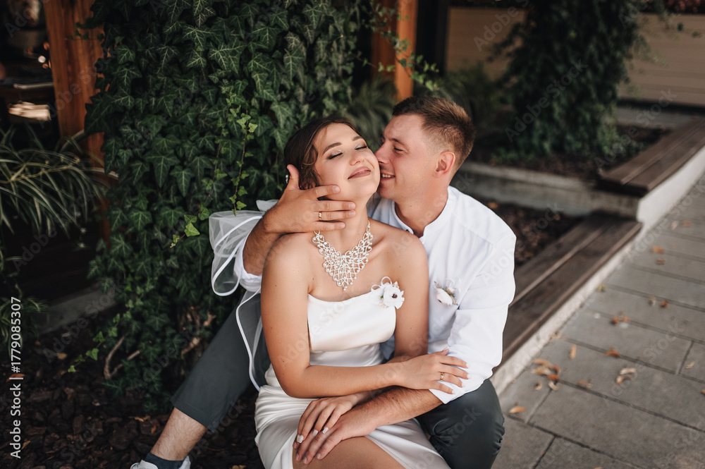 Fototapeta premium Wedding photography, portrait of happy smiling newlyweds, stylish groom and beautiful young brunette bride, hugging outdoors on the street in the city, in the park.
