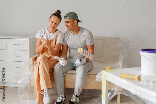 Two happy individuals sit on a couch surrounded by painting supplies, looking at their phones and enjoying their time together while preparing to redecorate