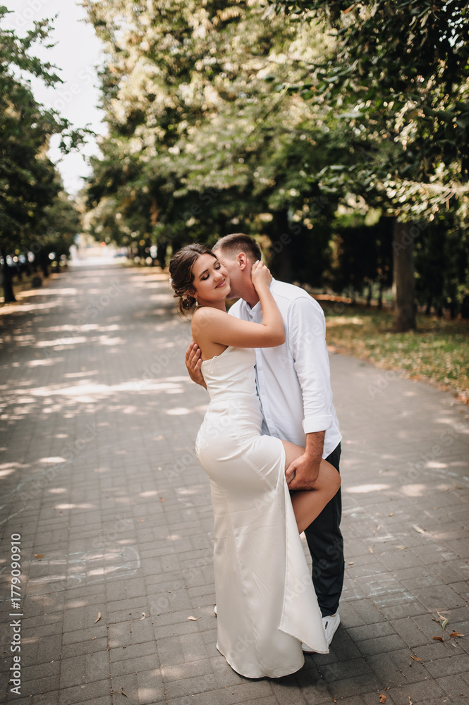 Naklejka premium Wedding photography, portrait of happy smiling newlyweds, a young groom and a beautiful sweet brunette bride, tenderly hugging outdoors on a street in the city, in a park.