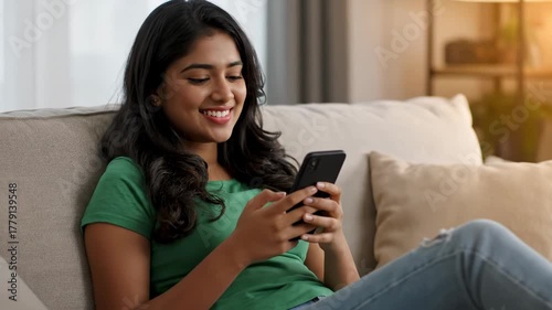 Young indian woman smiling while using her smartphone while relaxing on a couch at home