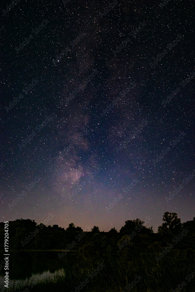 Fototapeta premium Stunning night landscape with the Milky Way stretching across the starry sky above a calm lake and dark forest silhouettes