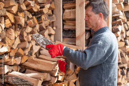 A man in red gloves picking up firewood from a large pile. An adult man in a gray sweater picks up firewood, against the backdrop of a large, neatly stacked woodpile