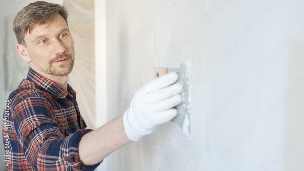 Fototapeta premium Male construction worker spreading plaster on white wall, wearing protective gloves during home renovation project with putty knife
