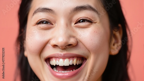 Young Asian woman laughing with perfect white teeth and glowing healthy skin on a soft coral background.