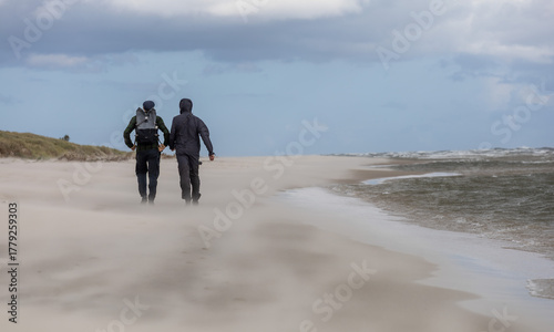 Fototapeta Naklejka Na Ścianę i Meble -  Two people walking along a windy beach beside the Baltic Sea, with sand blowing across the shore and waves rolling in under a cloudy sky in southern Sweden.