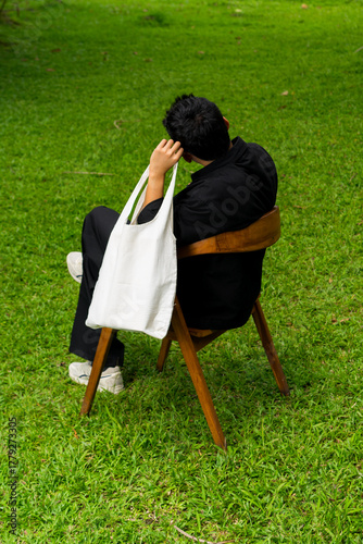 Man sitting on a wooden chair in the garden carrying a white tote bag on shoulder, symbolizing sustainable fashion and relaxed eco lifestyle.
