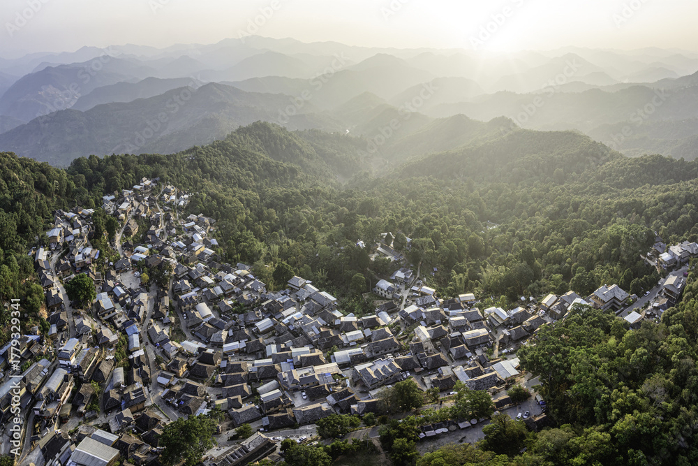© AmazingAerialAgency - Aerial view of a village nestled in lush green hills, with houses tightly packed together, contrasting with the distant hazy mountain ranges, Menghong Village, Yunnan, China.