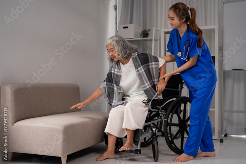 Asian nurse assisting older adult woman transfer from wheelchair to sofa providing dedicated support for home care patient mobility in rehabilitation