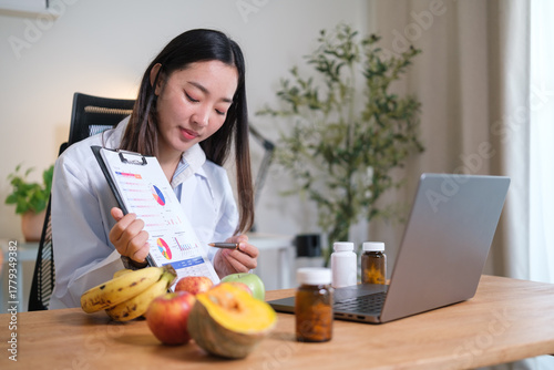 Female nutritionist explains a daily nutrition overview remotely, using data visuals while bananas, lettuce, carrots, pumpkin, and apples sit on the desk.