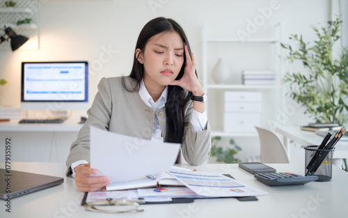 Fotografie Young businesswoman rubbing her temple while reading financial documents at a messy desk—calculator, charts, and paperwork—office stress concept