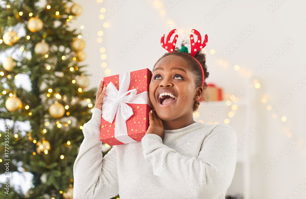 Naklejka premium Portrait of a happy African American woman laughing while holding a gift box, celebrating New Year or Christmas at home. Expresses amazement and joy as she enjoys the surprise or present.