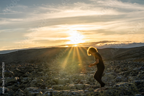 A cute little boy walks through sunbeams in the rocky mountains of Sweden. The setting sun casts a warm glow, creating a magical atmosphere during dusk.
