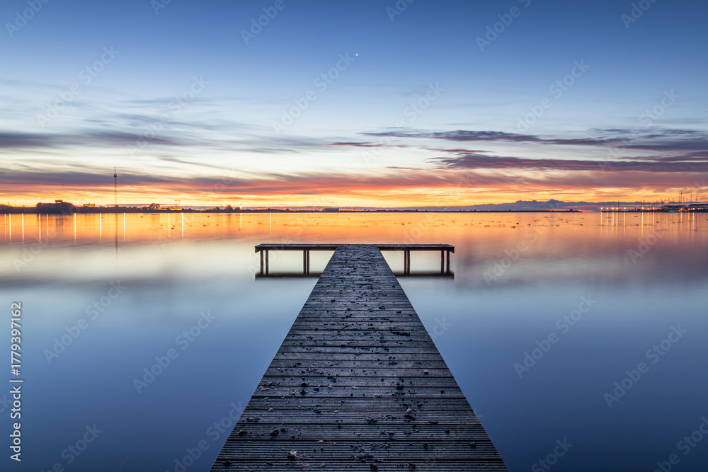 Naklejka premium Wooden pier in the Zuiderhaven of Den Oever during a colorful sunrise. The sun announces its arrival by painting the horizon orange, while the visible star remains as a reminder of the night.