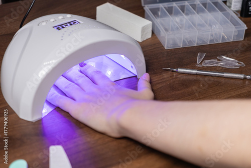 Woman's hand curing glitter purple gel nails in a modern LED UV lamp, performing a personal self-manicure beauty treatment