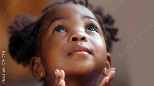 A close-up portrait of a cute African American girl praying, with her hands clasped and her eyes open, looking up, studio shot style
