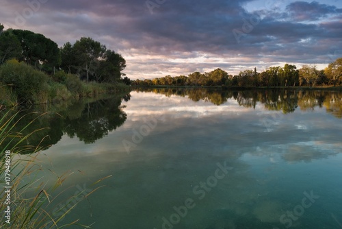 Fototapeta Naklejka Na Ścianę i Meble -  Tranquil lake reflecting colorful clouds under evening sky surrounded by lush greenery