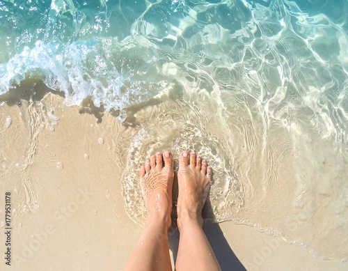 Close up of bare feet standing on a sandy beach with clear ocean water washing over them