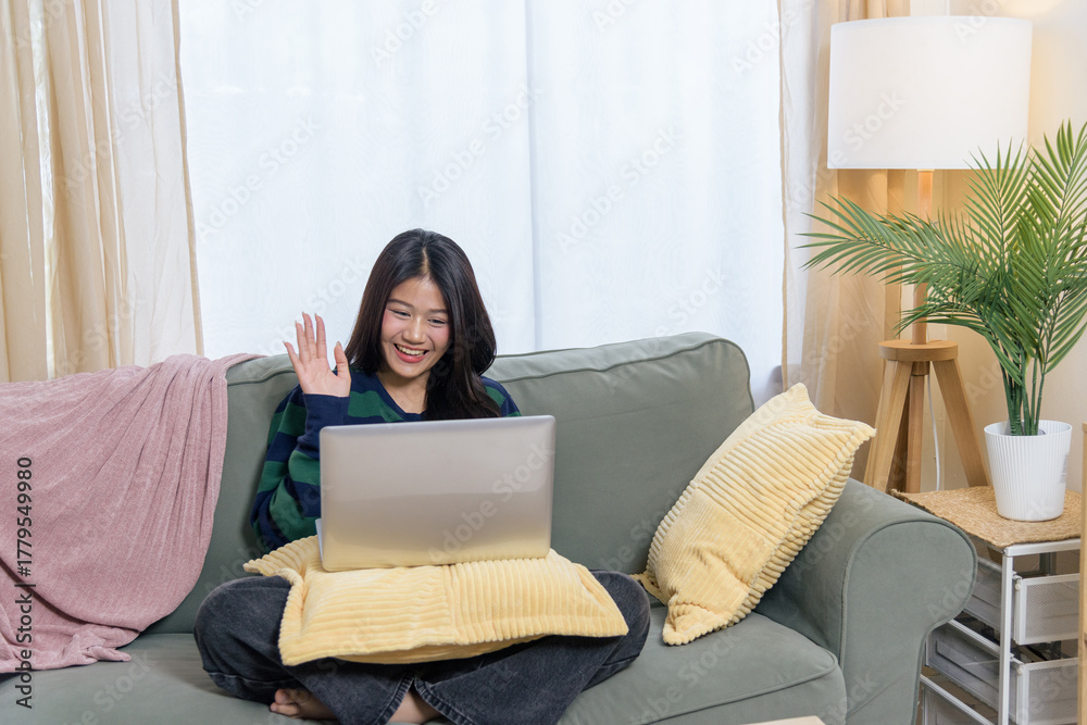 Fototapeta premium Happy teen woman relaxing at home, uses laptop for virtual meeting, waving hand, comfortable sofa supports remote work or online shop customer use.