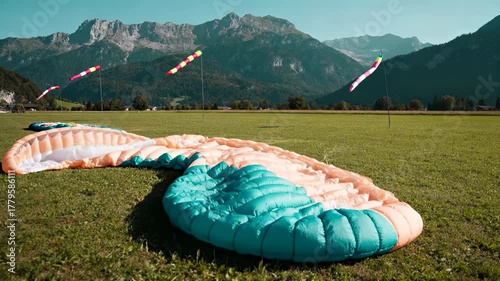 Large paraglider wing laid out on a green grassy field with wind socks and rugged Alps mountains in the background on a sunny day