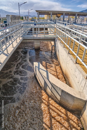 WWTP Aeration Tank: intense aeration of wastewater and activated sludge. Abundant brownish foam, concrete channels, and railings.