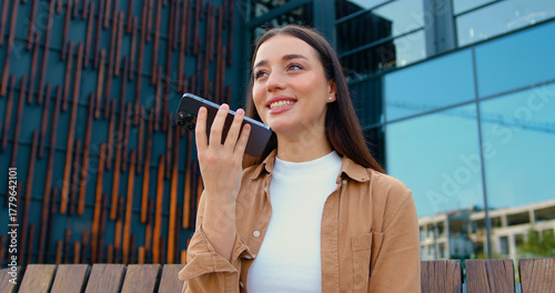 Young woman holding in hand smart phone talking with friend distantly uses easy voice messaging, concept of modern Ai technology, voice recognition, online translator over modern building background