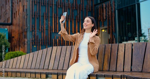 Close up of smiling Caucasian businesswoman talking on video call using phone outdoors while sitting on bench near office building. Online chat