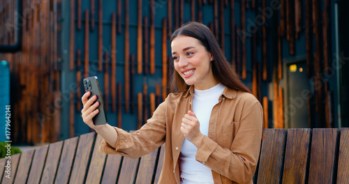 Close up of pretty Caucasian girl talking on video call using phone outdoors while sitting on bench near office building. Online chat