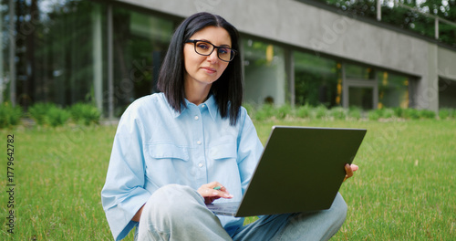 Portrait of a positive young woman sitting on grass in an urban park, using a laptop for remote work or study. Female student learning outdoors in a city garden