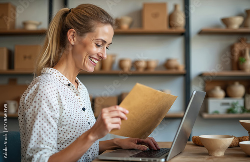 Happy woman in pottery workshop works on laptop holding package. Smiling female potter uses computer for online orders. Lady in ceramics studio smiles while at work © miss irine