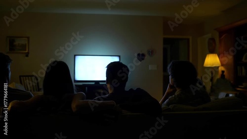 Group of people watching television together on a sofa in a dim living room at night, faces silhouetted by the screen glow, cozy movie night and streaming entertainment, family or friends bonding