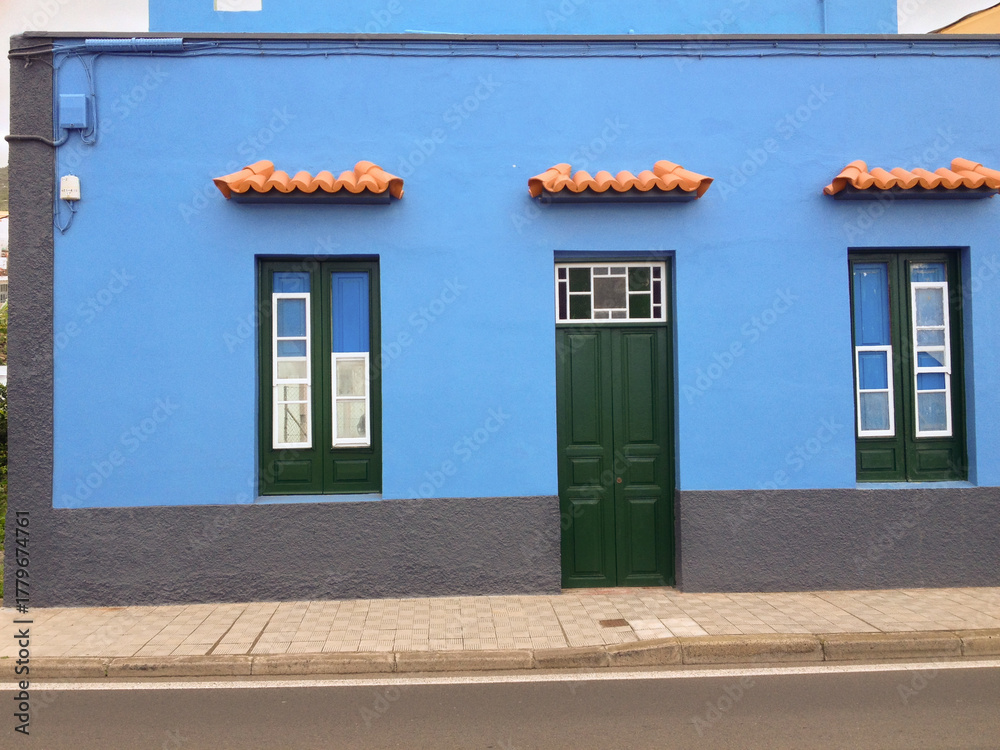 Naklejka premium Colorful blue house with green doors in Tenerife. Canary architectural style, bright contrast, and Mediterranean coastal charm.