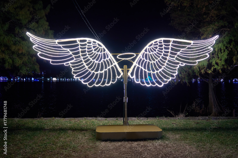 Fototapeta premium A glowing LED angel wings sculpture shines at night by the lake. Bright white lights create a festive holiday photo spot.