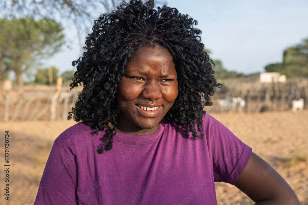 Fototapeta premium young african woman, in the village, at the farm in the yard, goats in the pen, sandy ground