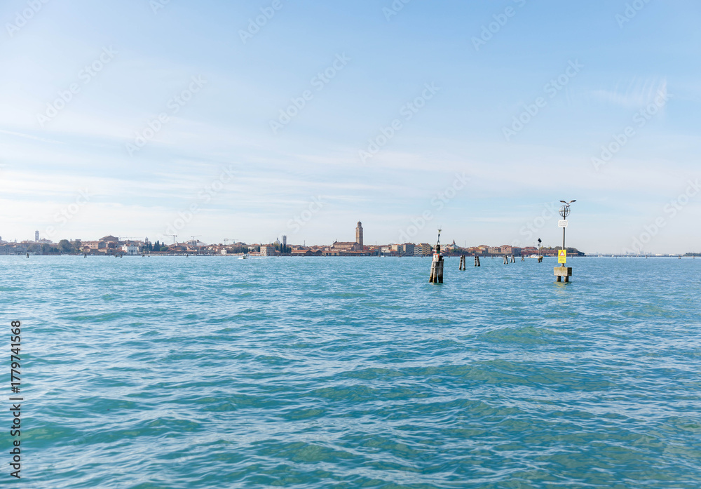 Fototapeta premium Murano, Italy - November 21, 2024: Scenic view of Murano Island with clear blue waters and distant buildings, showcasing the beauty of Venetian architecture and tranquil atmosphere