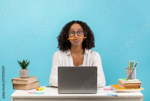 Fotografie A young black female student sits at a desk with books and a laptop, amusing herself by balancing a pencil on her face against a blue backdrop