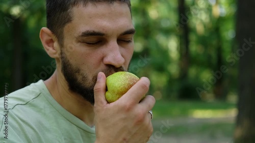 Close up of young man in light green t shirt eating fresh pear outdoors surrounded by green trees in sunny park. 4k horizontal footage