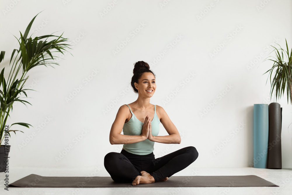 Fototapeta premium A calm smiling millennial woman practices meditation on a fitness mat in a bright studio. She sits in lotus position with clasped hands, surrounded by plants and yoga equipment.