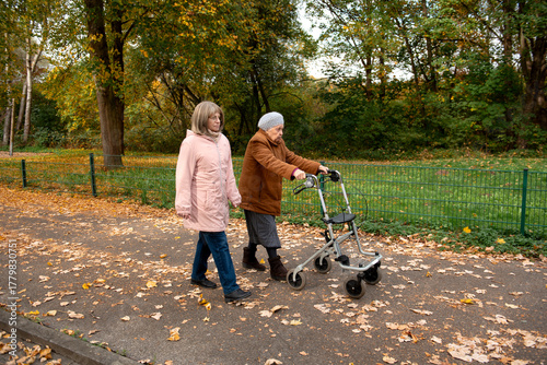 Woman and her elderly mother with walker taking a walk in the park covered in autumn leaves, spending family time together