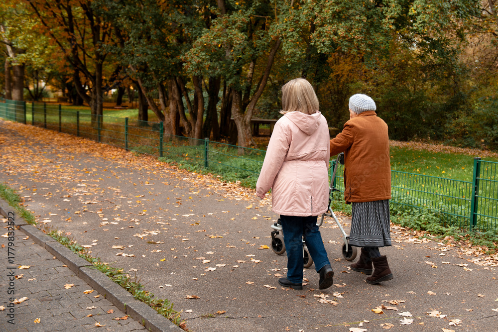 Fototapeta premium Woman and her elderly mother with walker taking a walk in the park covered in autumn leaves, spending family time together