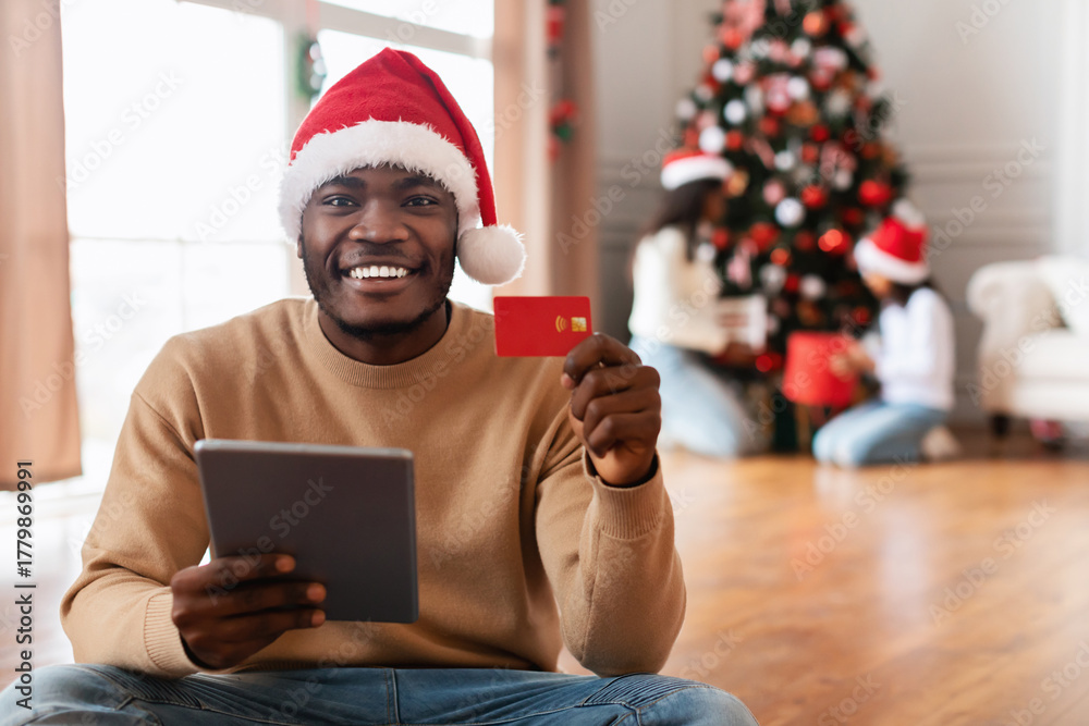 Fototapeta premium A happy man wearing a red Santa hat is sitting on the floor using a tablet and holding a credit card. In the background, his family is joyfully decorating a Christmas tree.