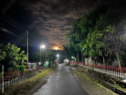 Quiet village street at night with glowing lamps and cloudy sky, creating a calm tropical mood.